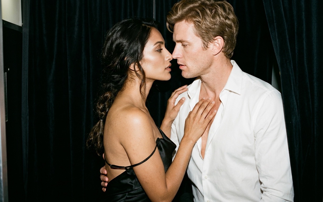 A South Asian woman with long dark hair in a loose braid, wearing a black satin slip dress with one strap fallen off her shoulder, stands with her hand at the open collar of a fair-skinned, sandy-haired man in a white shirt unbuttoned at the chest, his arm around her bare back, their faces close and not quite touching, lit by a hard white flash against a dark velvet curtain.