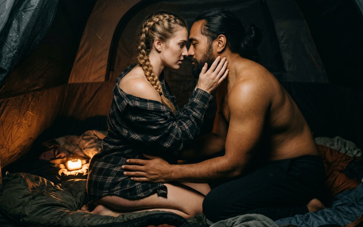 A woman with a honey-blonde braid, her plaid flannel shirt open and slipping off one bare shoulder, cups the jaw of a bare-chested, bearded man with his hair tied back as they kneel face to face inside a tent, his hand at her waist, a lit headlamp glowing beside the open sleeping bags around them.