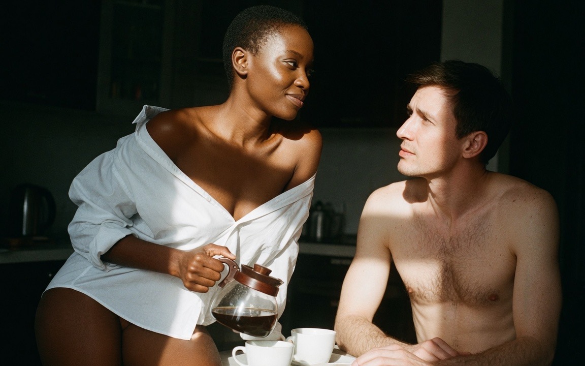 A woman in a man's loose white shirt slipping off one shoulder, holding a glass coffee pot over two white cups on a table and looking at a bare-chested man seated across from her, hard morning light falling across them and the kitchen dark behind.