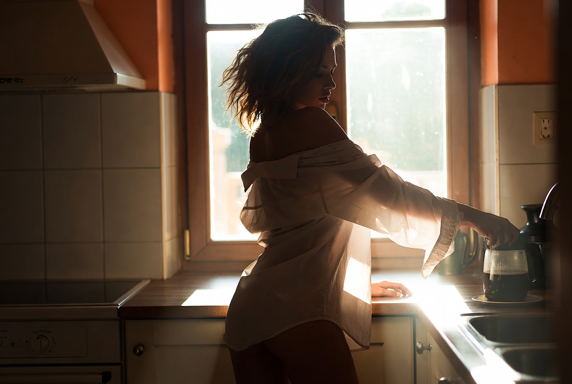 In a sunlit kitchen, a woman wearing only a man’s loose white shirt, slipped off one shoulder, reaches for a glass coffee pot on the counter, silhouetted against a bright window.