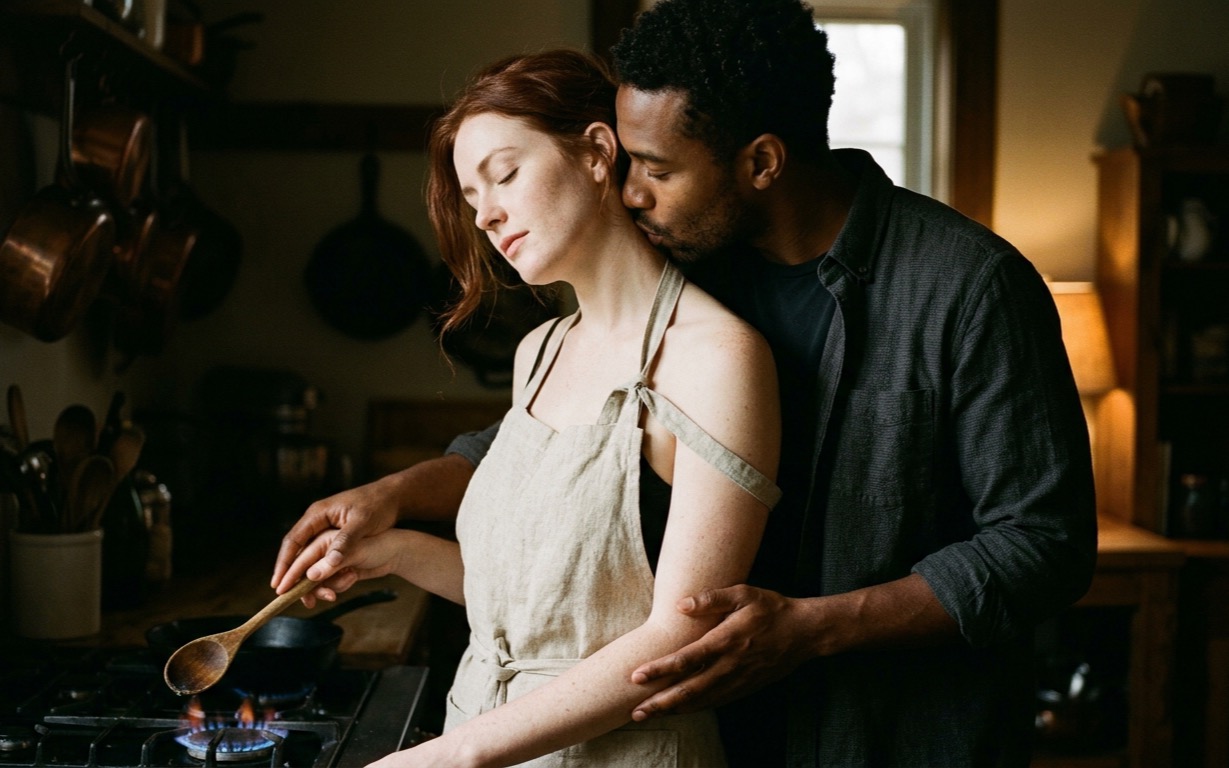 A red-haired, freckled woman in a linen apron with one strap slipped off her bare shoulder stands at a stove holding a wooden spoon over a pan on a low blue gas flame, eyes closed and head tipped back against a Black man behind her in a dark open shirt, his mouth at her neck and his hand resting over her forearm, copper pots hanging in the dim kitchen behind them.