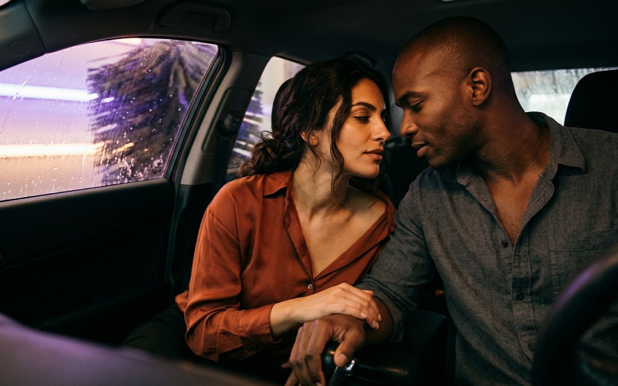 A woman with dark wavy hair in an unbuttoned rust-colored silk blouse sits in a car's passenger seat, her hand resting over a shaved-headed man's hand on the gearshift, their faces close and not touching, soap and violet-lit car-wash lights streaming across the window behind them.