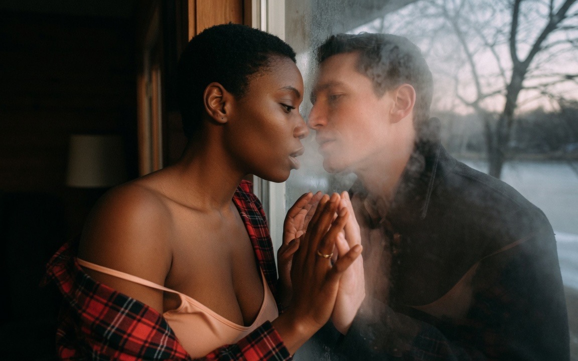 A woman in a red flannel shirt slipping off one shoulder over a peach slip, standing inside a lake house with her hands pressed together against a window pane, a man on the far side of the glass with his hands mirroring hers, their faces inches apart through the fogged window, a frozen lake and bare trees beyond him at dusk.