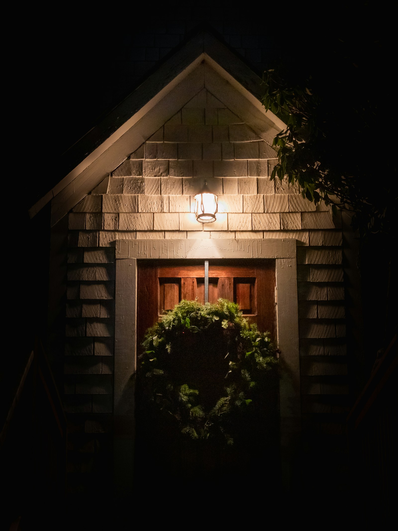 A single porch light glowing above a wreathed doorway, the rest of the house dark against the night.