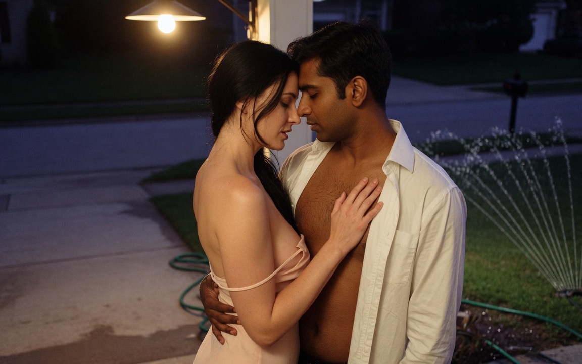 A woman in a pale slip dress with one strap fallen, turned into the chest of a man in an open white shirt on a suburban driveway at night, her hand flat on his chest and his arm around her waist, their foreheads together, a porch light above them and a lawn sprinkler running behind.