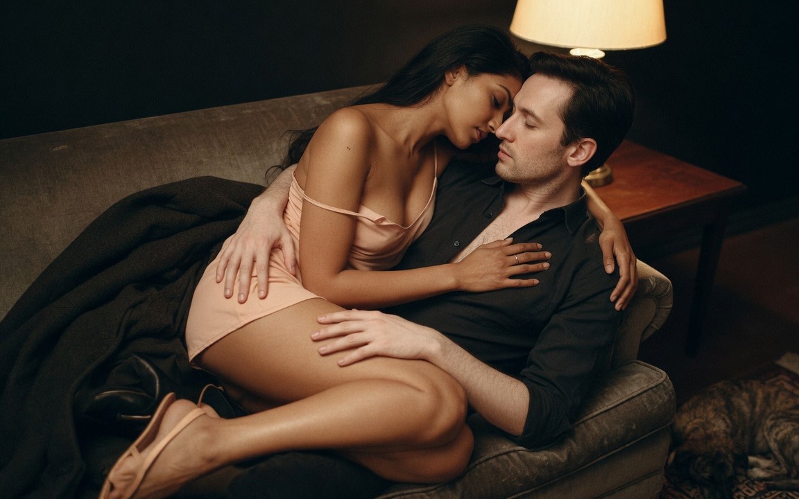 A woman in a pale pink slip curled sideways across a man's lap on a dark sofa, one strap fallen off her shoulder, her forehead resting against his cheek, her hand flat on his chest and his palm on her bare thigh, a wool coat dropped beside them and a lit lamp behind.