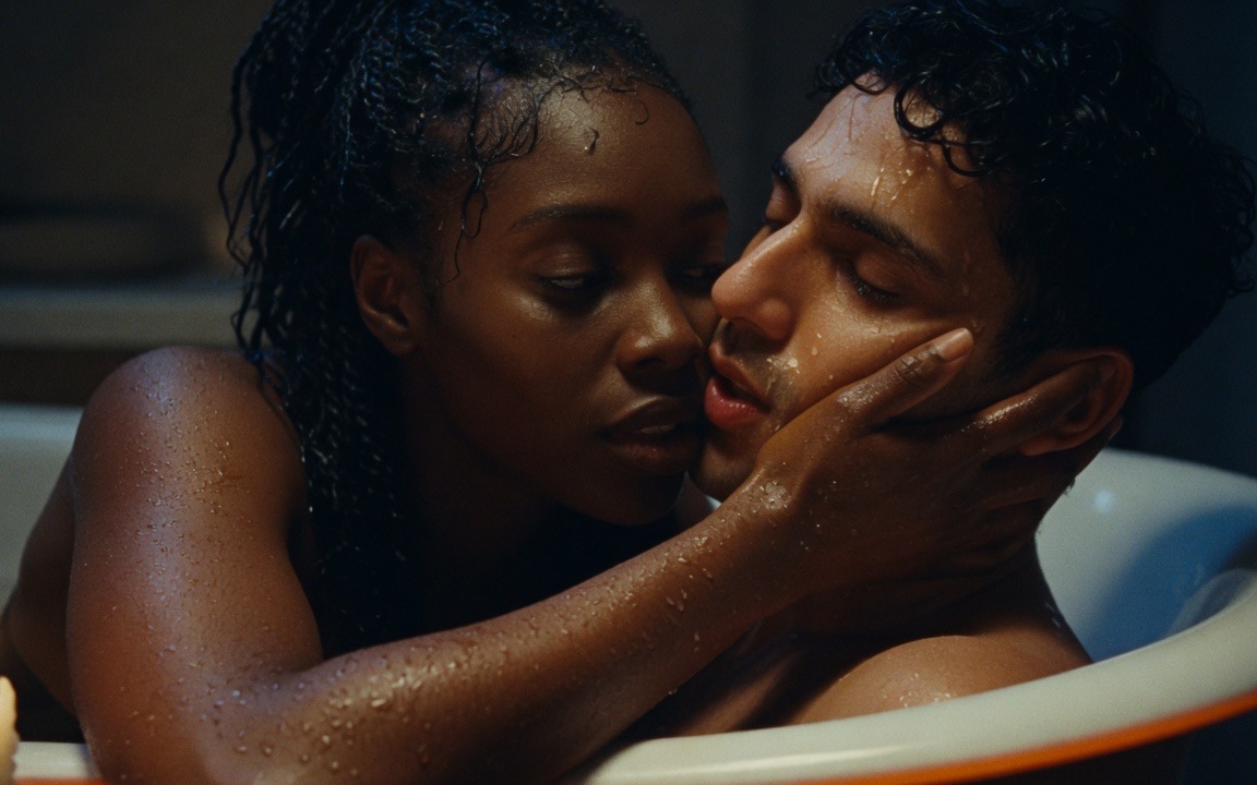 A close view of two people in a candlelit bath with wet skin and wet hair, a woman with long braids resting her hand along the jaw of a man whose face is turned into her palm with his eyes closed, the white rim of the tub across the lower edge of the frame.