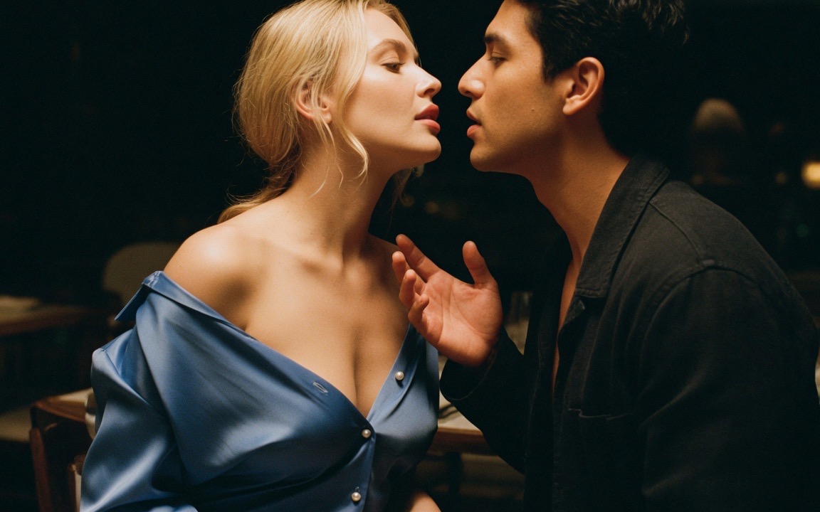 A woman in a blue silk dress slipped off both shoulders and fastened by three pearl buttons down the front, standing very close to a man in a dark jacket whose raised hand stops just short of touching her, their faces almost meeting, a dark restaurant behind them.