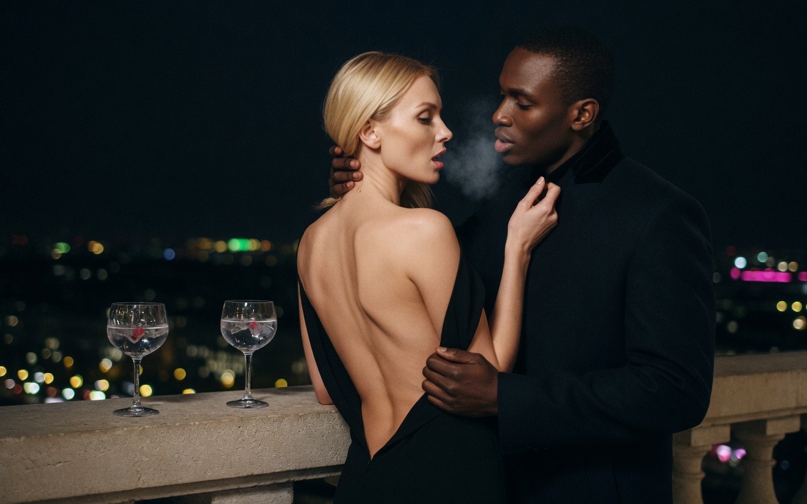A woman in a black evening dress cut open down her bare back turning toward a man in a dark overcoat on a high terrace at night, his hand at the back of her neck and her hand at his collar, their breath visible in the cold, two gin glasses on the stone rail and city lights far below.