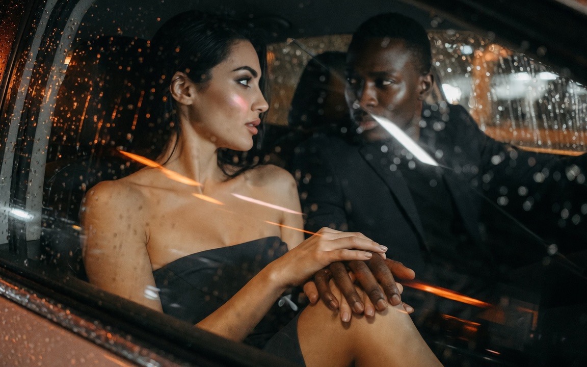 Seen through rain-streaked glass, a woman in a strapless dark dress sitting in a car at night beside a man in a dark suit, his hand resting on her bare knee and her hands laid over it, her face turned away toward the window, streaks of orange city light across the wet glass.