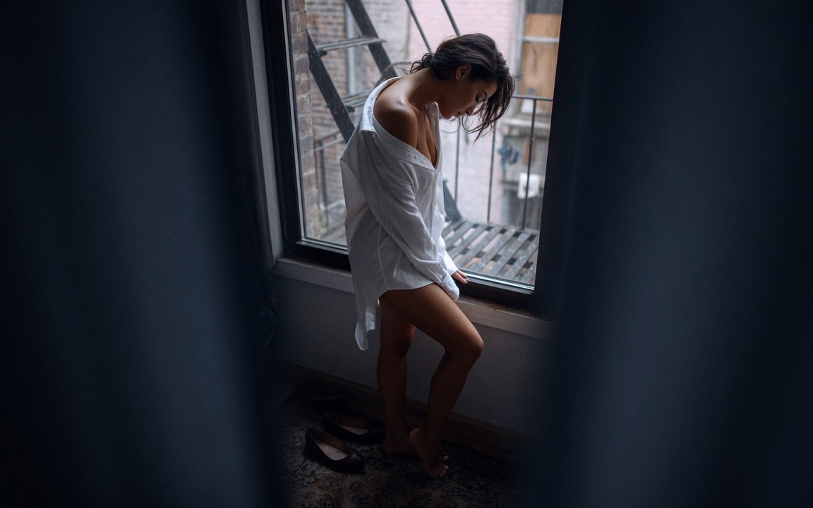 A woman in an oversized white shirt slipping off one shoulder, barefoot with bare legs, standing at a window with her head bowed, a fire escape and brick alley beyond the glass, her shoes set down on the rug beside her, seen between dark curtains.