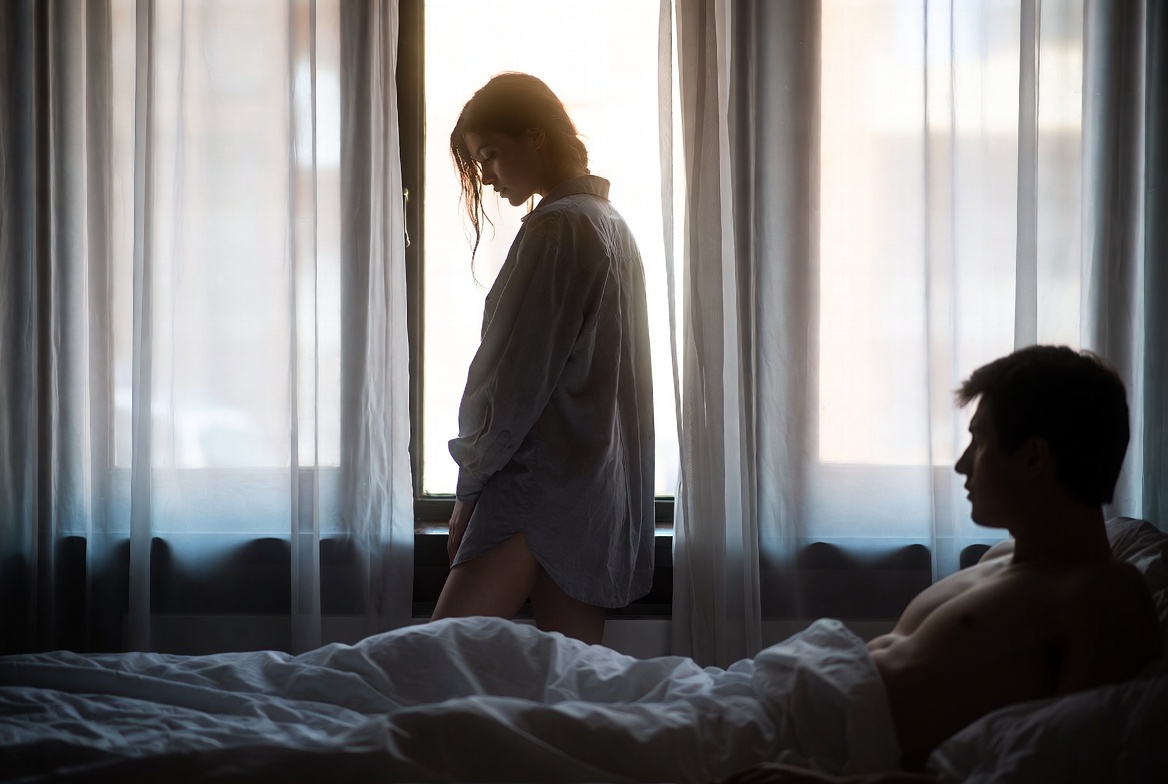 In gray morning light, a woman in an oversized button-down shirt stands at a curtained window with her head bowed, while a man watches her from the rumpled bed in the foreground.