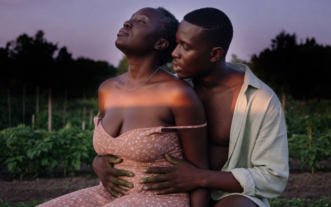 A Black woman in a pink floral sundress with the straps loosened at her shoulders, head tilted back against a bare-chested Black man in an open linen shirt behind her, his earth-stained hands resting at her waist, his mouth near her cheek, in a dusk garden with rows of green fading into shadow behind them.